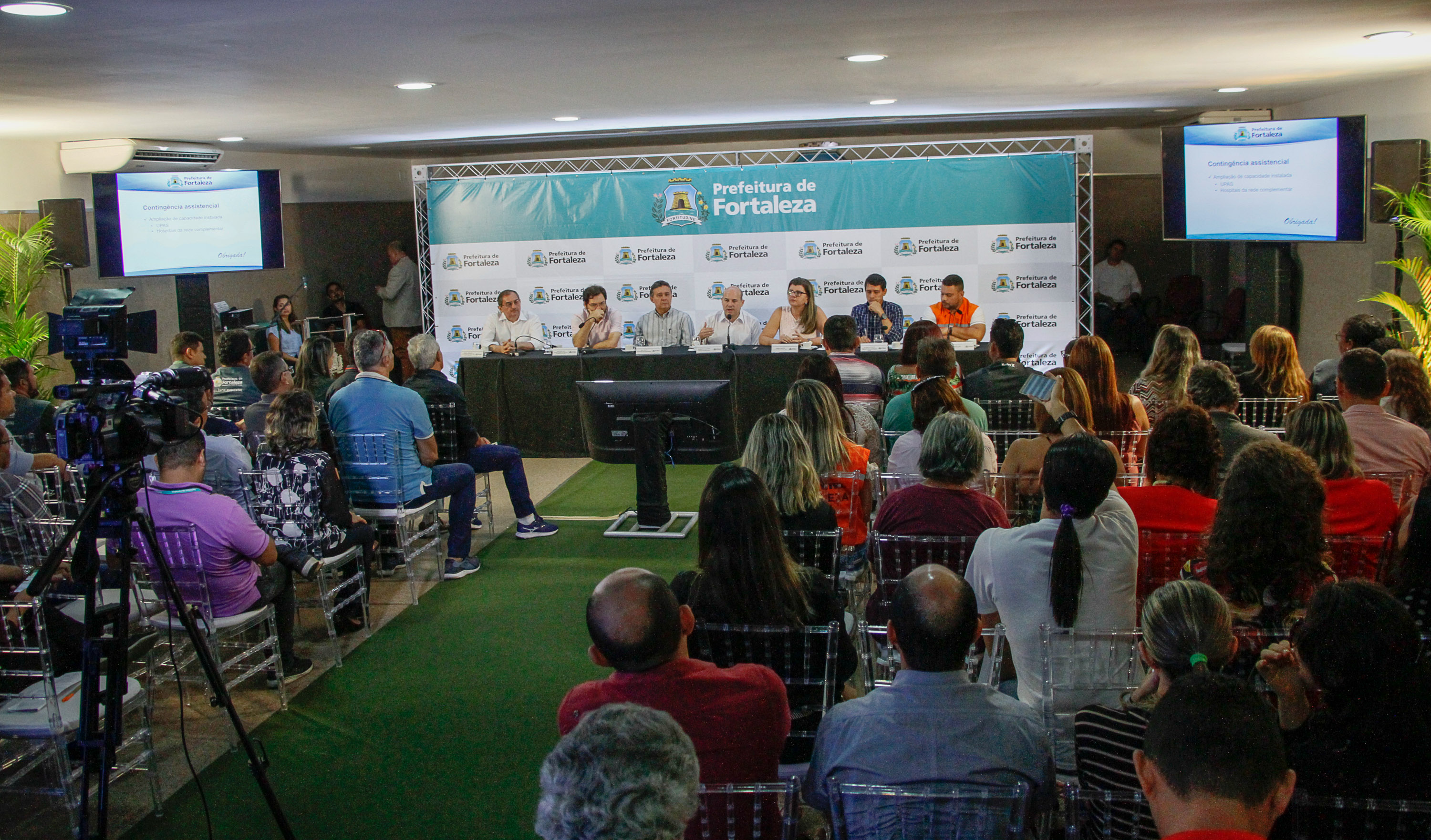 prefeito sentado à mesa falando ao microfone e demais pessoas sentadas ao lado e jornalistas sentados em frente assistindo